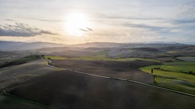 Gün batımında Val d 'Orcia' nın dalgalı tepelerini gösteren insansız hava aracı panoraması. Sıcak güneş ışığı tarlaları ve yeşil çayırları aydınlatarak tarım arazilerinin doğal dokusunu ve karakteristik selvi ağaçlarını aydınlatır.