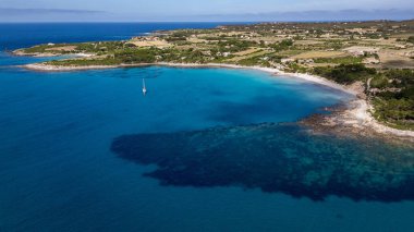Carloforte, İtalya 'daki Guidi Sahili' nin çarpıcı yüksek açılı manzarası. Görüntü, Sardunya kıyı şeridinin vahşi güzelliğini, kristal berrak turkuaz sularda demirli bir yelkenliyle, yoğun Akdeniz çalılıkları ve beyaz kumlu bir bataklıkla yakalıyor.