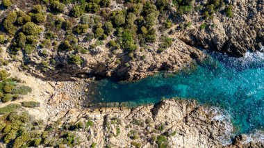 Güney Sardunya 'daki Cala Cipolla' nın el değmemiş sahil güzelliğini gösteren baş döndürücü bir hava fotoğrafı. Görüntü, yoğun Medi tarafından çerçevelenmiş şeffaf turkuaz deniz ile engebeli granit kayalıklar arasındaki canlı kontrastı vurguluyor.