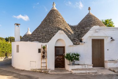 Alberobello, Puglia 'daki Trulli evleri. Beyazlatılmış duvarlar, konik taş çatılar, tencereli ahşap merdiven ve girişte beyaz çiçekler. UNESCO sitesi, Akdeniz kırsal mimarisinin başyapıtı. İtalya, Itria Vadisi.