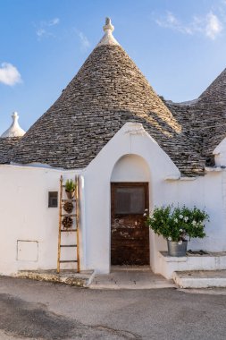 Alberobello, Puglia 'daki trullo girişinin ayrıntıları. Ahşap kapı, dekoratif merdiven ve çiçeklerle bembeyaz bir cephe. Mavi gökyüzünün altında tepe noktası olan konik taş çatı. UNESCO kırsal mimarisi, İtalya. Geleneksel Akdeniz tarzı.