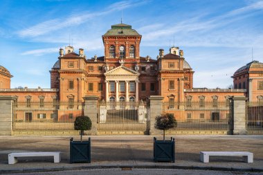 Racconigi Kalesi 'nin güney cephesinin simetrik görüntüsü. Barok ve Neoklasik kırmızı tuğla mimarisi, UNESCO Savoy konutu, önplanda berrak bir gökyüzünün altında anıtsal bir kapı ve modern banklar yer alıyor..