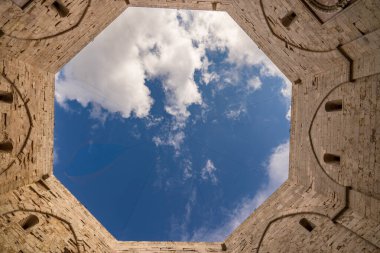 Castel del Monte 'nin sekizgen avlusuna bakıyorum. Kireçtaşı duvarlar geometrik olarak beyaz bulutlu mavi bir gökyüzü çerçeveler. Frederick II 'nin ortaçağ mimarisinin mükemmel simetrisi, Güney İtalya' nın Puglia kentindeki UNESCO Dünya Mirası.