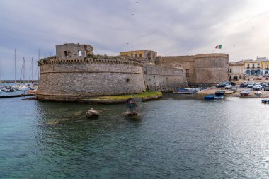Rivellino kulesi ve denizden yükselen Urchin heykeliyle Gelibolu Kalesi manzarası. Limana demirlemiş tekneler ve bulutlu bir gökyüzünün altında tarihi Salento mimarisi. Güney İtalya, Apulia 'nın ikonik kıyı manzarası.