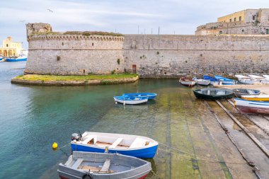 Kristal berrak denizden yükselen görkemli Rivellino kulesiyle Gelibolu Kalesi manzarası. Küçük mavi ve beyaz tekneler bulutlu bir gökyüzünün altında tarihi duvarlar boyunca demirlemiştir. Salento, Apulia, Güney İtalya 'nın ikonik kıyı manzarası.