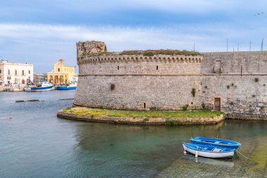 Limana demirlemiş devasa taş duvarları ve mavi tekneleriyle Gelibolu Kalesi manzarası. Arka planda, bulutlu bir gökyüzünün altındaki Salento köyünün tarihi mimarisi. Güney İtalya, Apulia 'nın ikonik ve turist kıyı manzarası.
