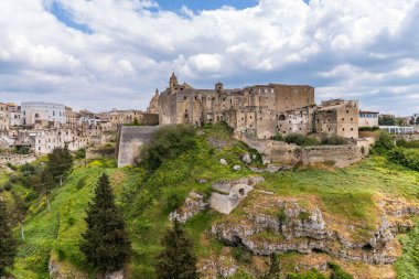 Puglia 'daki Gravina manzaralı derin vadiye bakan taş evler. Arka planda, Akdeniz yeşillikleri arasındaki antik viyadük köprüsü dramatik bulutlu bir gökyüzünün altında. Iconic kayalık arazisi ve İtalya 'daki antik köy.