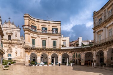 Martina Franca 'daki Piazza Maria Immacolata manzarası. Fotoğrafta Barok yarı dairesel Portikolar, zarif ışıklı taş binalar ve arka planda San Martino Bazilikası, Puglia 'nın Itria Vadisi' nin kalbi yer alıyor.