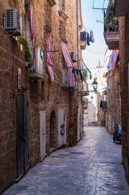 Taranto 'nun eski kasabasındaki dar bir sokak manzarası. Uzun carparo taşından binalar, çamaşır asma, çizgili tenteler ve park halindeki Goril Piaggio bu tarihi güney İtalya şehrinin kalbinde otantik ve zamansız bir atmosfer yaratıyor..