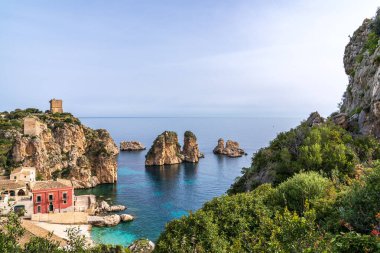 Sicilya 'daki tarihi Tonnara di Scopello' nun panoramik manzarası. Kırmızı bina ve taş yapılar kristal berrak sudan yükselen deniz yığınlarını gözden kaçırıyor. Antik gözetleme kuleleri açık bir gökyüzünün altındaki kayalıklara hükmediyor. İkonik İtalyan kıyı manzarası.