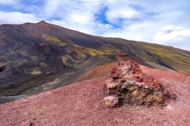 Etna Dağı 'nın Silvestri Kraterleri' ndeki gözenekli kırmızı lav kayasına yakın çekim. Kırmızı volkanik toprak, İtalya 'nın Sicilya kentindeki Avrupa' nın en yüksek aktif yanardağının karmaşık jeolojisini gözler önüne seren yeşil yamaçlar ve bulutlu gökyüzü ile çelişiyor. Manzara Görünümü.