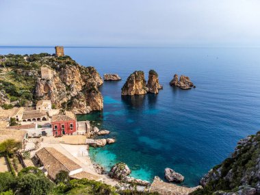 Sicilya 'daki tarihi Tonnara di Scopello' nun panoramik manzarası. Kırmızı bina ve taş yapılar kristal berrak sudan yükselen deniz yığınlarını gözden kaçırıyor. Antik gözetleme kuleleri açık bir gökyüzünün altındaki kayalıklara hükmediyor. İkonik İtalyan kıyı manzarası.