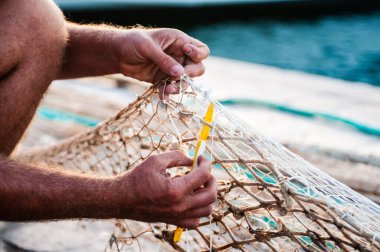 Geleneksel bir balıkçının yetenekli ellerine yakın çekim. Sarı ağ iğnesiyle beyaz bir ağı onarıyor. Görüntü denizcilik geleneğini ve günlük ağır işçiliğin sabrını bir rıhtımda ya da liman ortamında yakalıyor..
