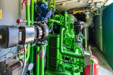Bir biyogaz tesisinin içindeki yeşil endüstriyel yanmalı motorun detaylı görüntüsü. Fotoğrafta, yenilenebilir enerji teknolojisi ve sürdürülebilir enerji üretimini temsil eden kompleks borular, vanalar, elektrik pompaları ve kırmızı basınç tankı yer alıyor..