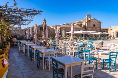 Marzamemi 'deki Regina Margherita Meydanı manzarası. Önde beyaz masalar, mavi sandalyeler, sarı kaplar ve bir pergola var. Arka planda, San Francesco di Paola Kilisesi Sicilya, İtalya 'da güneşli bir günde açık mavi bir gökyüzünün altında duruyor..