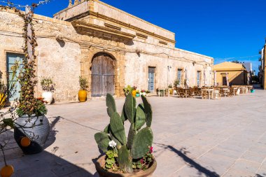 Marzamemi 'deki Regina Margherita Meydanı manzarası. Ön planda dikenli bir armut kaktüsü var. Arka plan Villadorata Sarayı 'nın taş cephesini gösteriyor. Geniş ahşap bir kapısı, açık kafe masaları ve İtalya' da açık mavi ve güneşli bir gökyüzü var..