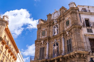 Palermo 'daki Quattro Canti' nin görkemli Barok cephesinin alçak açılı görüntüsü. Resim, kralların ve patronların heykellerini, dövülmüş demir balkonları ve parlak mavi bulutlu gökyüzüne karşı zengin taş süslemeleri vurguluyor..