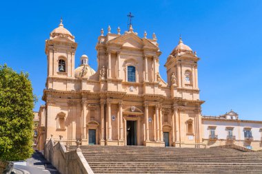 İki kule tarafından çerçevelenen ve Bernardo Labisi 'nin ikonik merdiveni tarafından desteklenen Noto Katedrali' nin görkemli cephesinin alçak açılı görüntüsü. Açık mavi gökyüzünün altındaki altın kireç taşı detayları. İtalya 'nın Sicilya kentindeki UNESCO Dünya Mirası.
