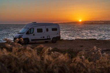 Sicilya 'daki Punta Pisciotto uçurumunda parketmiş beyaz bir minibüsün anımsatıcı görüntüsü. Görüntü, Akdeniz üzerinde sıcak altın gün batımını ön planda yabani otlar ve huzurlu bir özgürlük atmosferiyle yakalıyor..