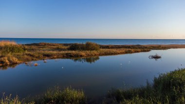 Sicilya 'daki Belice Doğa Rezervi' nin panoramik görüntüsü. Sakin bir göl, kumlu kum tepeleri ve Akdeniz çalılıklarıyla denizden ayrılan mavi gökyüzünü yansıtır. Güney İtalya 'da barış ve biyolojik çeşitliliğin el değmemiş vahası..