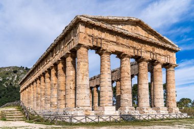 Segesta Doric Tapınağı 'nın açısal perspektifi. Altın taş yapı sisli mavi bir gökyüzüne karşı duruyor. Ön planda, ahşap bir çit ve taş basamaklar Sicilya Arkeoloji Parkı 'na giden yolu gösteriyor..