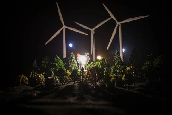 Electricity power in nature or clean energy concept. Wind Turbine producing alternative energy at night. Glowing bulb powered by alternative energy. Creative decoration with small miniature. Selective focus