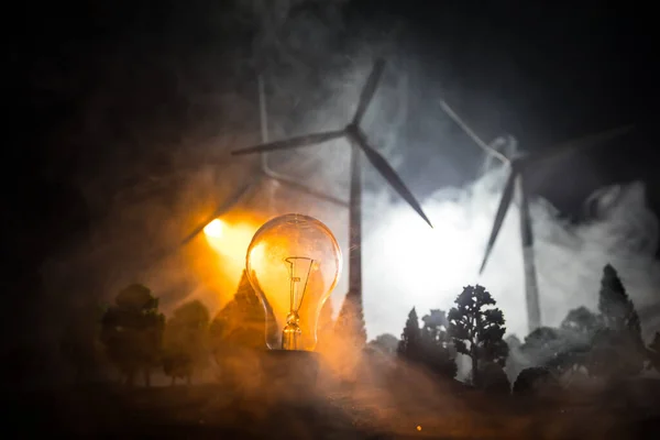 Electricity power in nature or clean energy concept. Wind Turbine producing alternative energy at night. Glowing bulb powered by alternative energy. Creative decoration with small miniature. Selective focus