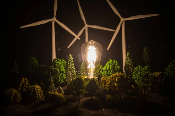 Electricity power in nature or clean energy concept. Wind Turbine producing alternative energy at night. Glowing bulb powered by alternative energy. Creative decoration with small miniature. Selective focus