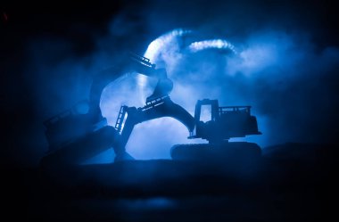 Şehir caddesinde bir inşaat alanı. Gece boyunca inşaat alanında park etmiş sarı kazıcı kazıcısı. Karanlık sisli arka planda endüstriyel konsept masa dekorasyonu. Seçici odak