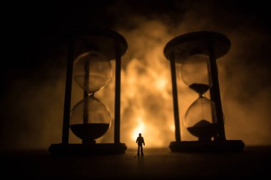 Zaman kavramı. Kum saatleri arasında duran, dumanlı ve karanlık arka planda ışıkları olan bir adamın silueti. Gerçeküstü dekore edilmiş resim