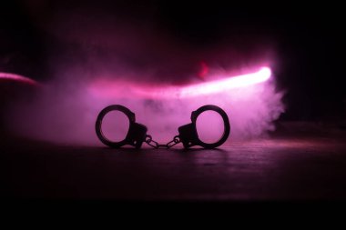 Gece polis baskını ve sen tutuklusun. Arka tarafında polis arabası olan kelepçelerin silueti. Görüntü sisli arka planda yanıp sönen kırmızı ve mavi polis ışıkları. Seçici odak