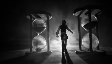 Zaman kavramı. Kum saatleri arasında duran, dumanlı ve karanlık arka planda ışıkları olan bir adamın silueti. Gerçeküstü dekore edilmiş resim