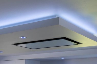 Çağdaş bir mutfak tavanında hafif mavi LED ışıklandırması ve yansıtıcı cam yüzeyi olan floş montajlı bir kaput bulunur.