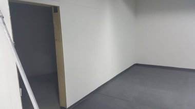 Beyaz duvarları olan çıplak bir iç mekan, karanlık bir iç mekanı gösteren açık bir kapı, ve siyah süpürgelikli koyu gri bir kat.