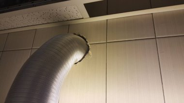 Gümüş esnek bir havalandırma hortumu bej kaplama duvardaki bir delikten geçirilir ve tavan paneli görünür hale gelir.