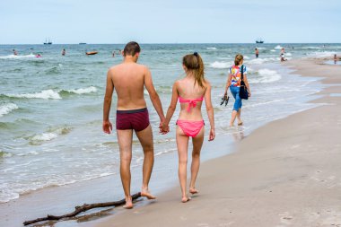 LEBA, POLAND - 31 Temmuz 2016: Yazın sahilde yürüyen genç bir çift. Erkek arkadaşıyla el ele tutuşan bikinili bir kadın. Deniz kıyısında yürü.