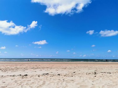 Geniş kumlu sahil, mavi gökyüzü, beyaz bulutlar ve güneşli bir yaz gününde okyanus ufku. Seyahat, tatil ve rahatlama kavramları için geniş bir kopya alanına sahip doğal kıyı manzarası.