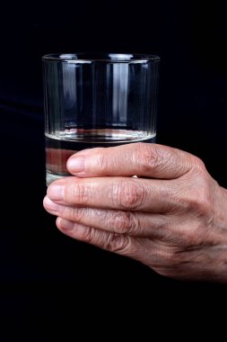 Yaşlı bir kadının kırışık ellerinde bir bardak su. Siyah arka planda yakın plan.