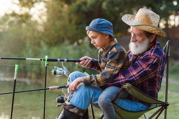 Sympathisch respektierter 70-jähriger bärtiger Großvater, der seinen 10-jährigen niedlichen Enkel auf Knien hält und ihm beibringt, Fische zu fangen. — Stockfoto
