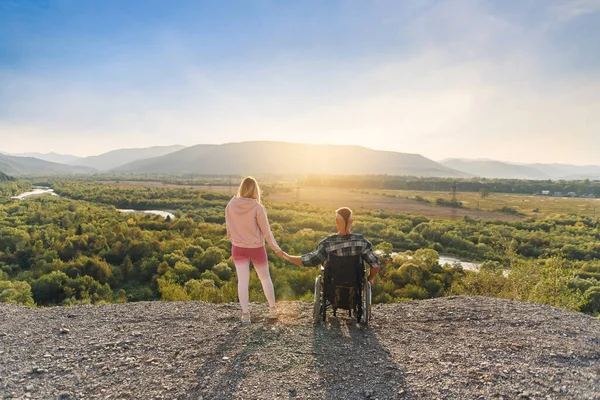Beautiful woman and her disabled husband in wheelchair holding hands on a mountain hill. — Stock Photo, Image