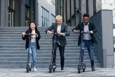 Eğlenceli, çok ırklı ofis arkadaşları dışarıda modern ofis yakınlarında elektrikli scooterlarda yarış yapıyorlar..