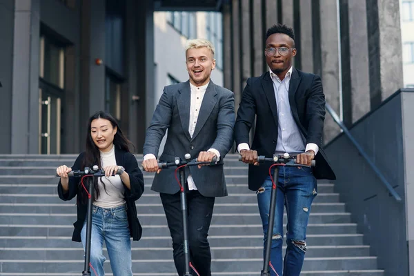 Divertidos colegas de oficina multirraciales alegres que tienen competencia de carreras en scooters eléctricos cerca de la oficina moderna fuera. — Foto de Stock