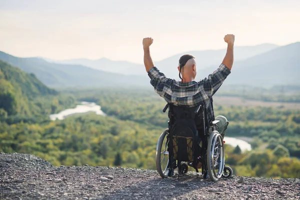 Неповносправний хлопець сидить у інвалідному кріслі з руками вгору на горі в оточених красивих пейзажах — стокове фото