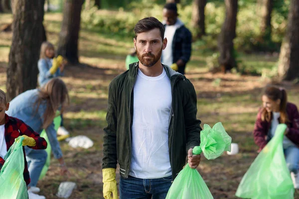 Good-looking bearded man shows a full rubbish pack in background of his friends volunteers picking up rubbish at park.