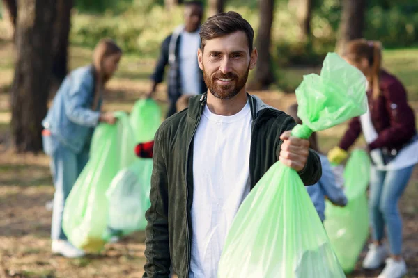 Handsome smiling bearded man holds rubbish bag in the background of his friends activists collecting rubbish at the park
