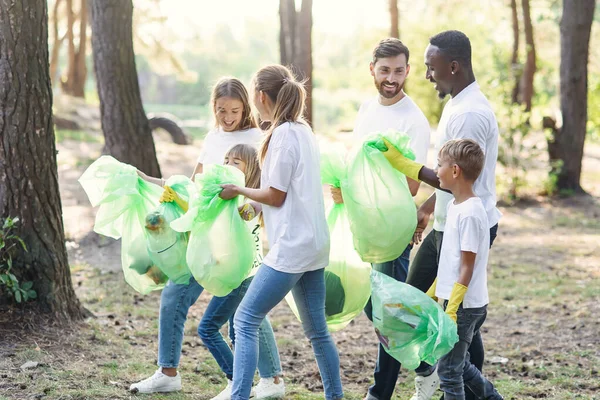 Активная команда любителей природы со всего мира в белых футболках собирает мусор в пластиковые пакеты в лесу. Лицензионные Стоковые Изображения
