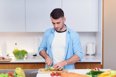 Yakışıklı adam sıcak mutfakta taze sebzelerden salata hazırlamak için ahşap bir tahtada domates kesiyor. Sağlıklı yemek..