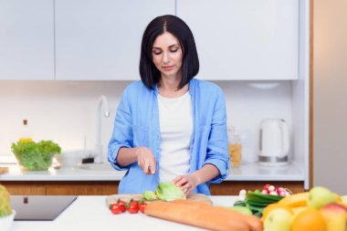 Genç bir kadın ahşap bir tahtanın üzerinde bıçakla lahana keser ve ev mutfağında sebzelerden salata hazırlar..