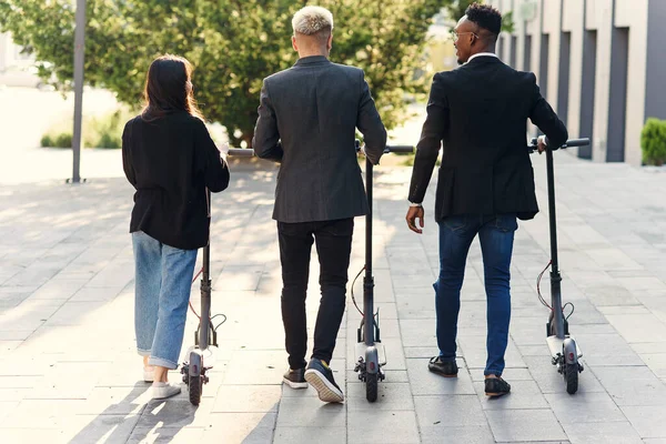 Colaboradores multiétnicos profesionales sostienen scooters eléctricos caminando cerca de un edificio moderno y hablando — Foto de Stock