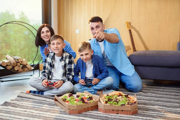 Pai feliz, mãe e dois filhos sentados no chão, jogando videogames com gamepads e comendo pizza saborosa. — Fotografia de Stock